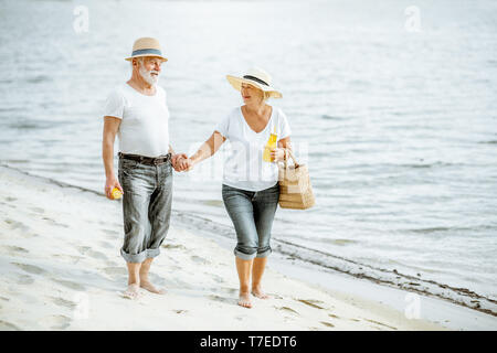 Felice coppia senior vestite di bianco t-shirt e cappelli a camminare insieme sulla spiaggia di sabbia durante il loro pensionamento Foto Stock