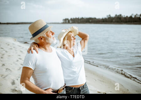 Felice coppia senior vestite di bianco t-shirt e cappelli insieme rilassante sulla spiaggia di sabbia durante il loro pensionamento Foto Stock