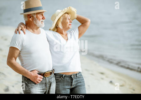 Felice coppia senior vestite di bianco t-shirt e cappelli a camminare insieme sulla spiaggia di sabbia durante il loro pensionamento Foto Stock