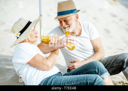 Felice coppia senior rilassante, giacente con bevande sulla spiaggia sabbiosa, godendo della loro pensione vicino al mare Foto Stock