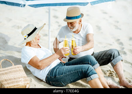 Felice coppia senior rilassante, giacente con bevande sulla spiaggia sabbiosa, godendo della loro pensione vicino al mare Foto Stock