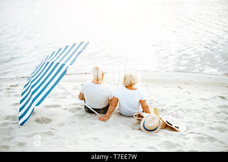 Bella coppia senior rilassante, seduti insieme con ombrellone sulla spiaggia sabbiosa, godendo della loro pensione vicino al mare, vista posteriore Foto Stock