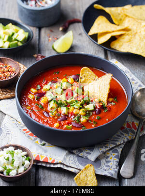 Pomodoro messicano, fagiolo, peperone zuppa nel recipiente nero con nachos chips. Sfondo di legno. Foto Stock