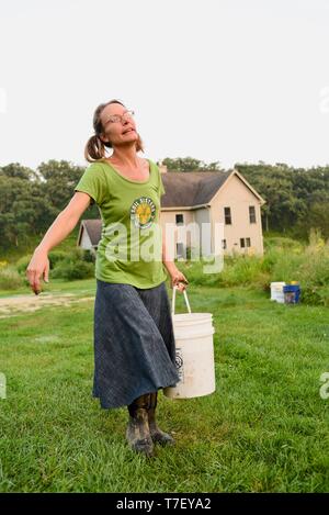 Donna agricoltore sul suo lavoro fattoria di maiale, alimentando il patrimonio di razza di porco con mangimi nel secchio, agriturismo in distanza, Blanchardville, WI, Stati Uniti d'America Foto Stock