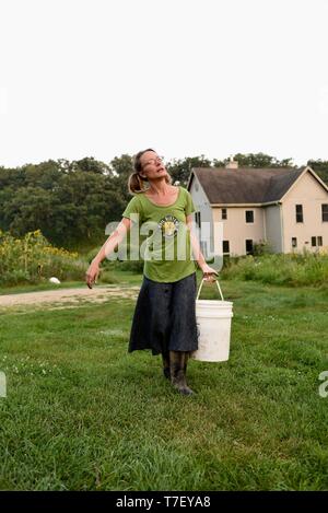 Donna agricoltore sul suo lavoro fattoria di maiale, alimentando il patrimonio di razza di porco con mangimi nel secchio, agriturismo in distanza, Blanchardville, WI, Stati Uniti d'America Foto Stock