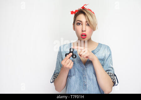 Hai spin? Ritratto di bella sorpresa giovane donna casual in denim blu con camicia, rosso permanente con fascia, tenendo spinner e puntamento figrer ad esso Foto Stock