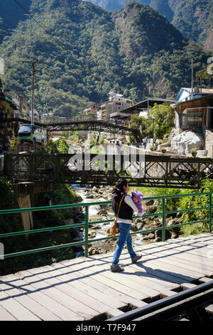 Donna Peruviana con il suo bambino camminando sul ponte sopra l'Aguas Calientes fiume di Machupicchu Puebo (noto anche come Aguas Calientes) in Perù Foto Stock