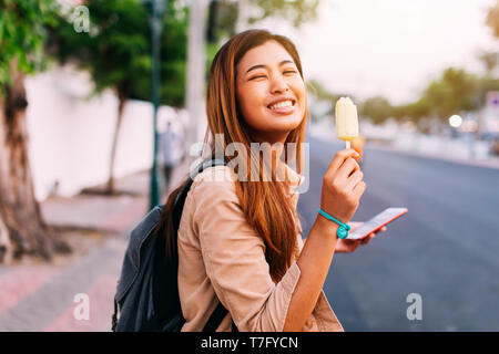 Vista laterale della incantevole donna asiatica tenendo lo smartphone e mangiare gelato mentre sorridente in telecamera sulla strada soleggiata Foto Stock
