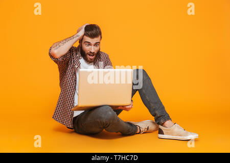 Ritratto di un scioccato giovane uomo che indossa abiti casual seduta isolate su sfondo giallo, utilizzando il computer portatile Foto Stock