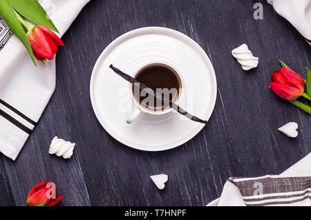 Nero caffè in una tazza bianca, il baccello di vaniglia, marshmallow, zucchero raffinato in forma di cuori e tulipani rossi Foto Stock