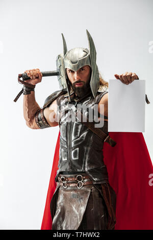 Dio del tuono. Bionda capelli lunghi e muscolare modello maschile in pelle Viking's costume con il grande martello di Thor cosplaying su bianco di sfondo per studio. Fantasy guerriero, il concetto di affari. Copia dello spazio. Foto Stock