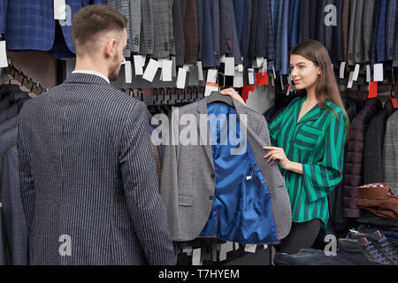 Piuttosto shop consulente scegliendo, azienda e mostrando giacca grigia per il giovane. Uomo che guarda un grande scelta di abiti e capi di abbigliamento maschile del quartiere alla moda di stampe su appendiabiti in fila. Foto Stock