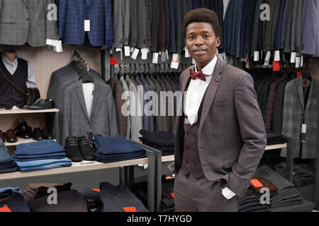 Client di boutique tenendo la mano in tasca dei pantaloni, guardando la fotocamera. Uomo bello il montaggio e posa in abito grigio, camicia bianca e rossa il filtro bow tie. Ampia scelta di abbigliamento moderno su appendiabiti e ripiani. Foto Stock