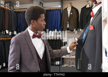 Giovane africano in piedi nella boutique di abbigliamento per uomini nei pressi di manichino, guardando alla camicia. Client di negozio in camicia bianca con red bow tie, nero waiscoat, giacca grigia scelta elegante abito. Foto Stock