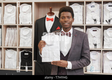 Giovane africano tenendo una camicia bianca e in posa nella boutique di abbigliamento uomo. Uomo in camicia bianca, rossa bow tie, gilet e giacca grigia guardando la fotocamera. Ripiani con magliette su sfondo. Foto Stock