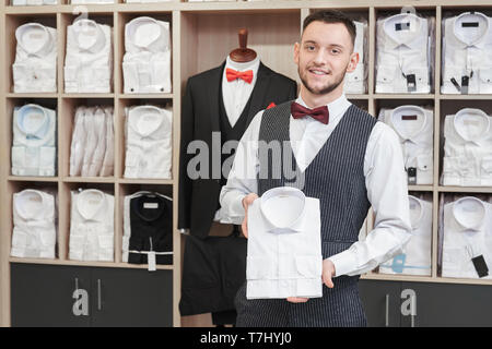 Ritratto di giovane uomo, client di boutique sullo sfondo con magliette su ripiani. Fiducioso uomo con barba tenendo una camicia bianca, guardando la telecamera, in posa. Boutique di moda per gli uomini. Foto Stock