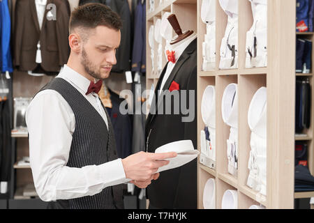 Giovane assistente con la barba scegliendo shirt su ripiani in boutique. Bello client in una camicia bianca con red bow tie, panciotto guardando i ripiani, camicia di contenimento. Abbigliamento elegante per gli uomini. Foto Stock