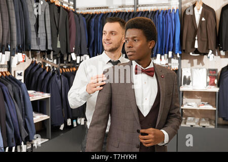 Assistant aiutando e scegliendo tuta per uomo africano nel negozio elegante. Client in una camicia bianca, gilet, giacca e red bow tie e venditore con la barba in piedi nel centro del negozio, guardando lontano. Foto Stock