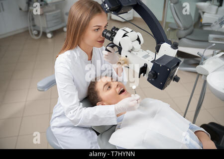Stomatologist esaminando denti del paziente con aiuto di professional microscopio dentale. Dentista prevenire la carie utilizzando strumenti di ripristino. Client giacente sulla poltrona del dentista. Foto Stock
