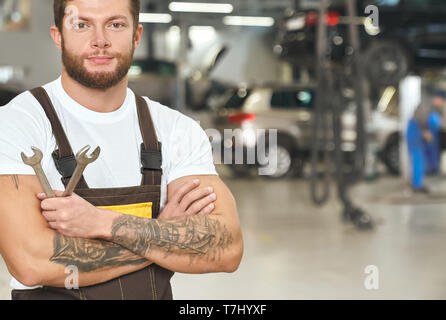 Brutali, tattoed repairman azienda chiavi, posa, guardando la fotocamera. Bello, muscolare uomo che indossa in bianco di t-shirt, tute, lavorando come meccanico nel moderno, professional autoservice. Foto Stock