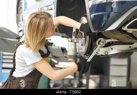 Vista laterale del meccanico femmina tenendo la chiave, riparare il fissaggio del disco del freno di automobile sollevato sul ponte. Giovane donna che indossa in uniforme lavora con veicolo in autoservice. Foto Stock