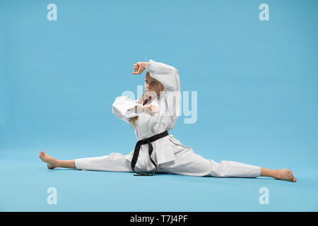 Forte bambina indossa un kimono bianco seduto su longitudinale dello spago e cercando di lato in studio. Bambino attivo in posa mentre la formazione blu su sfondo isolato. Concetto di arti marziali e karate. Foto Stock