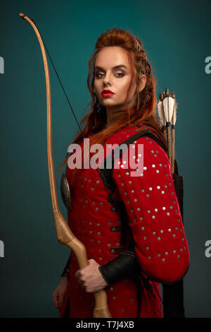 Splendida donna con capelli allo zenzero, labbra rosse guardando la telecamera, in posa. Bellissima femmina guerriero prua tenuta in mano, con frecce dietro la schiena, indossa in rosso tunica medievale. Foto Stock