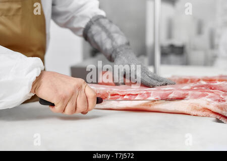 Chiusura del macellaio mani in guanti speciali il taglio di pezzi di carne fresca con il coltello. Tritare la carne di maiale o di carni bovine sul tavolo in macelleria. La produzione di carne. Industria alimentare Foto Stock