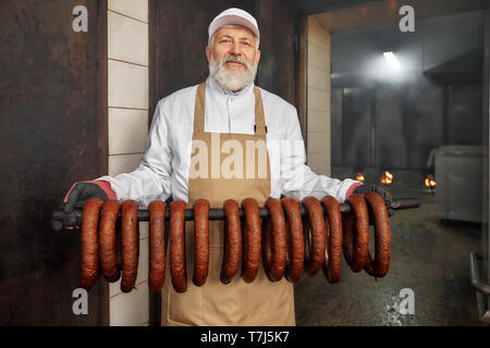 Produzione di insaccati. Anziani, bello lavoratore in piedi in smokehouse con salsicce affumicate, guardando la fotocamera. Sassages affumicato appeso in fila. Uomo in bianco uniforme, marrone grembiule, posa. Foto Stock