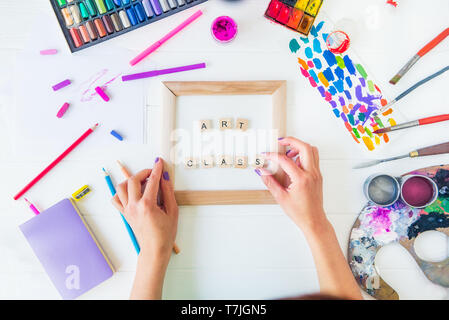 Vista superiore arte creativa il concetto di classe. Mani femminili aggiungere lettera di iscrizione sulla tela con molte colorate paintiing materiali su bianco backgroun in legno Foto Stock