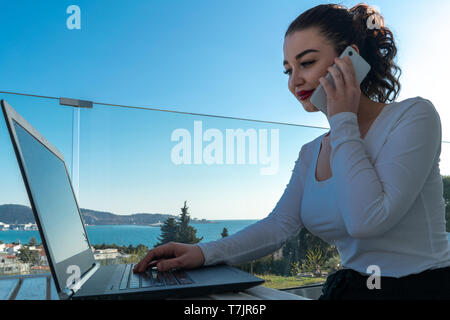 Donna attraente lavorando sul computer portatile sul balcone e godersi lo splendido paesaggio panorama. Vista sul mare, sulle montagne e la città. Freelance di luogo di lavoro. Conne Foto Stock