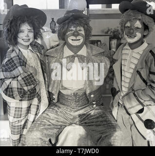 Anni sessanta, storico, un circo clown in visita ad una scuola elementare seduto accanto a con i giovani della scuola i bambini sia indossando abiti fantasiosi o abiti divertenti e con le loro facce dipinte come un clown, Inghilterra, Regno Unito. Foto Stock