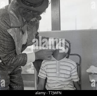 Anni sessanta, storico, un maschio circus clown visitare una scuola primaria messa clown faccia o vernice su un giovane scolaro seduto in una sedia, Londra, Inghilterra, Regno Unito. Foto Stock