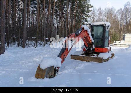 Un escavatore rosso nei pressi di una foresta è coperto da neve, come il sito di costruzione si sono inattivi a causa della bassa temperatura, neve e ghiaccio. Foto Stock