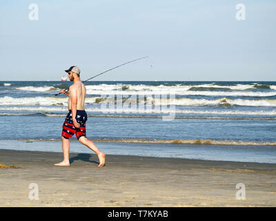 Un pescatore con esche penzolanti dalla pole in pantaloncini corti e tappo a sfera passeggiate sulla spiaggia accanto all'acqua in Port Aransas, Texas, Stati Uniti d'America. Foto Stock