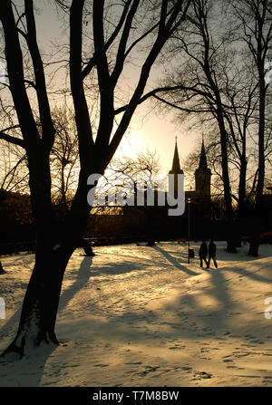 Due persone a piedi in un parco al tramonto a Tallinn in Estonia con St Charles' chiesa in background e gli alberi casting lunghe ombre sulla neve Foto Stock