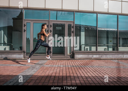 Una donna bella, una bambina corre in estate in città, su un Morning jog, pelle abbronzata. Sportswear leggings top. Gli auricolari per telefono. Concetto di fitness jogging Foto Stock