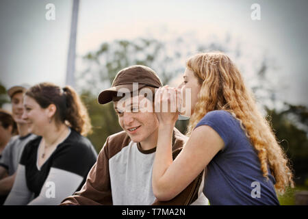 Ragazza adolescente whispering nel suo fidanzato l orecchio mentre seduto fuori con gli amici. Foto Stock