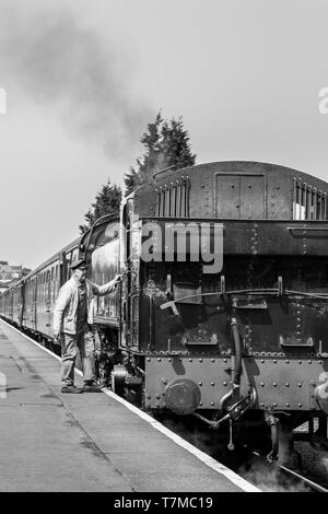 Pilota di equipaggio di treno a vapore bianco e nero, in berretto e tuta in fiocchi, su piattaforma ferroviaria che si imbarcarsi in attesa di una locomotiva a vapore britannica d'epoca, estate. Foto Stock