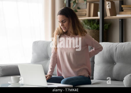 Donna seduta sul lettino vicino al computer portatile soffre di dolori alla schiena Foto Stock