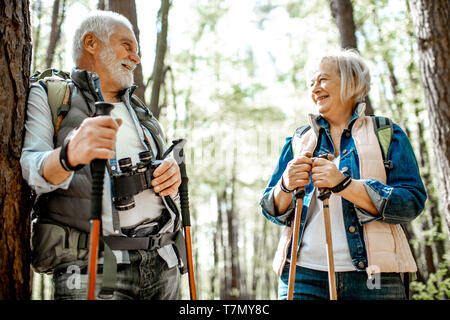 Coppia senior adagiata vicino l'albero mentre si viaggia con zaini e bastoncini da trekking nella foresta. Concetto di uno stile di vita attivo su pensionamento Foto Stock