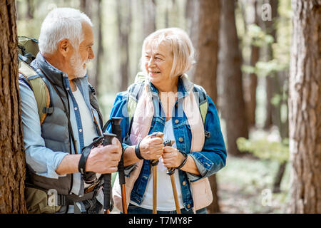 Coppia senior adagiata vicino l'albero mentre si viaggia con zaini e bastoncini da trekking nella foresta. Concetto di uno stile di vita attivo su pensionamento Foto Stock