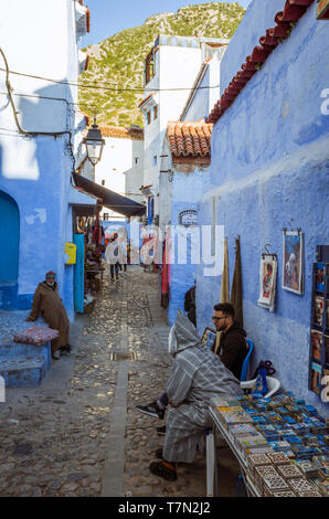 Chefchaouen, Marocco : uomini sedersi davanti a un negozio di souvenir si blocca su un vicoletto nel blu-lavato medina old town. Foto Stock