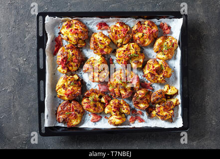 Australian Crash Hot per le patate di primizia cosparso con spezie e pancetta fritta e erbe su una teglia su una tavola di cemento, vista orizzontale dal di sopra, flatlay Foto Stock