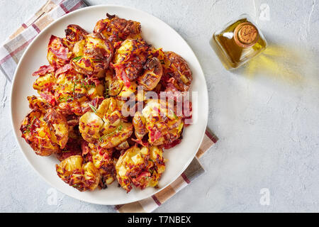 Close-up di Crash Hot per le patate di primizia cosparso con spezie e pancetta fritta e erbe su una piastra bianca su una tavola di cemento, cucina australiana, vista orizzontale Foto Stock