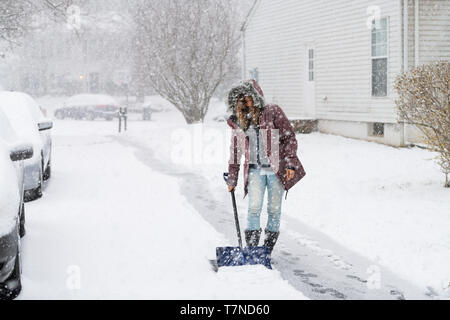 Giovane donna in cappotto invernale spalare la pulizia vialetto street dalla neve in pesante tempesta di neve con la pala da case residenziali e di automobili parcheggiate su strada Foto Stock