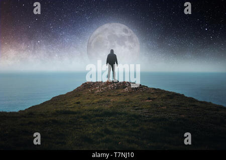 Un concetto di incappucciati figura in piedi su una collina che guarda al mare, con la luna e le stelle e l'universo di cui sopra. Foto Stock