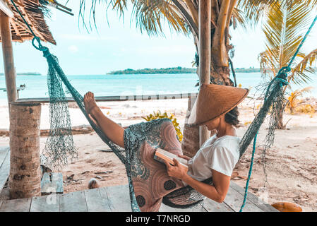 Donna che legge un libro sul amaca spiaggia tropicale, gente reale allontanarsi da tutto e da tutti, tradizionale South Asian hat, palme, teal dai toni arancione Foto Stock