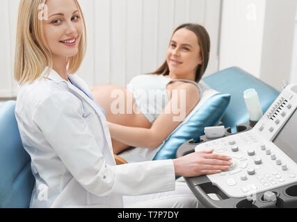 Vista dal lato della bella femmina medico guardando la fotocamera e sorridente durante la procedura in clinica moderna. Professional ginecologo facendo diagnostica ad ultrasuoni della madre durante la gravidanza. Concetto di medicina. Foto Stock