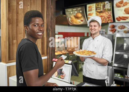 Client maschio guardando la fotocamera e sorridere mentre l'acquisto di una deliziosa pizza al cafe'. Ragazzo africano in piedi vicino a cashbox e pagando con carta di credito per posta indesiderata dood in pizzeria. Concetto di fast food e il pranzo. Foto Stock